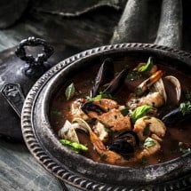 Cioppino Seafood Stew Soup in dark metal pot on gray wood surface.