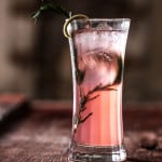 Tall glass filled with pink liquid and rosemary garnish in shadow.