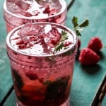 Two garnished glasses of homemade raspberry soda on green wood surface