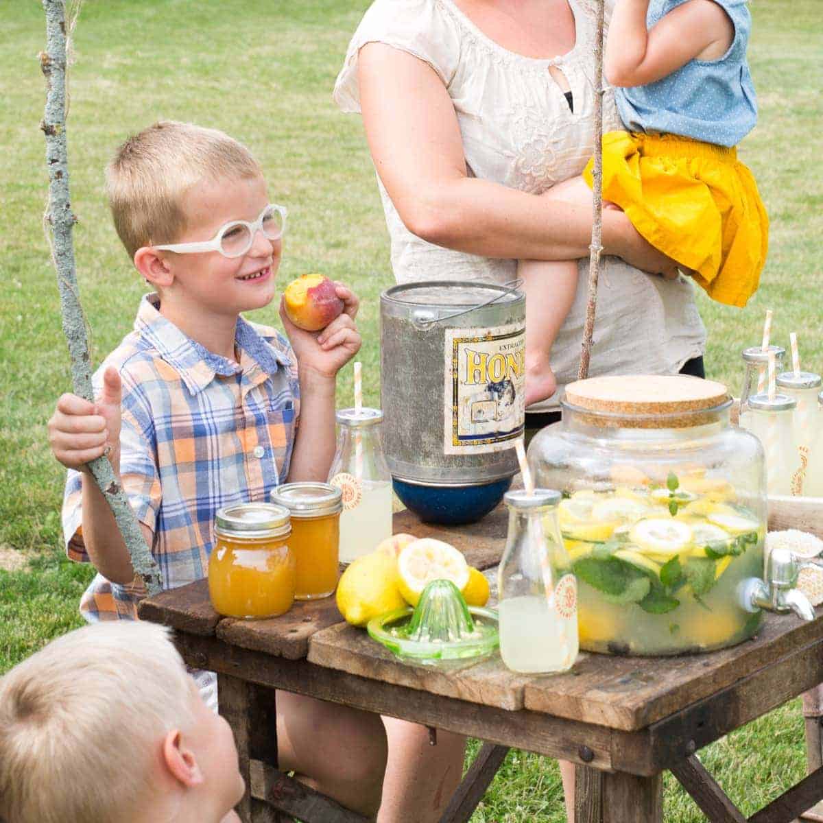 Boy in glasses smiling and eating a peach behind lemonade stand