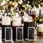 Three glass bottles of homemade syrup with chalkboard signs in front of a Christmas tree