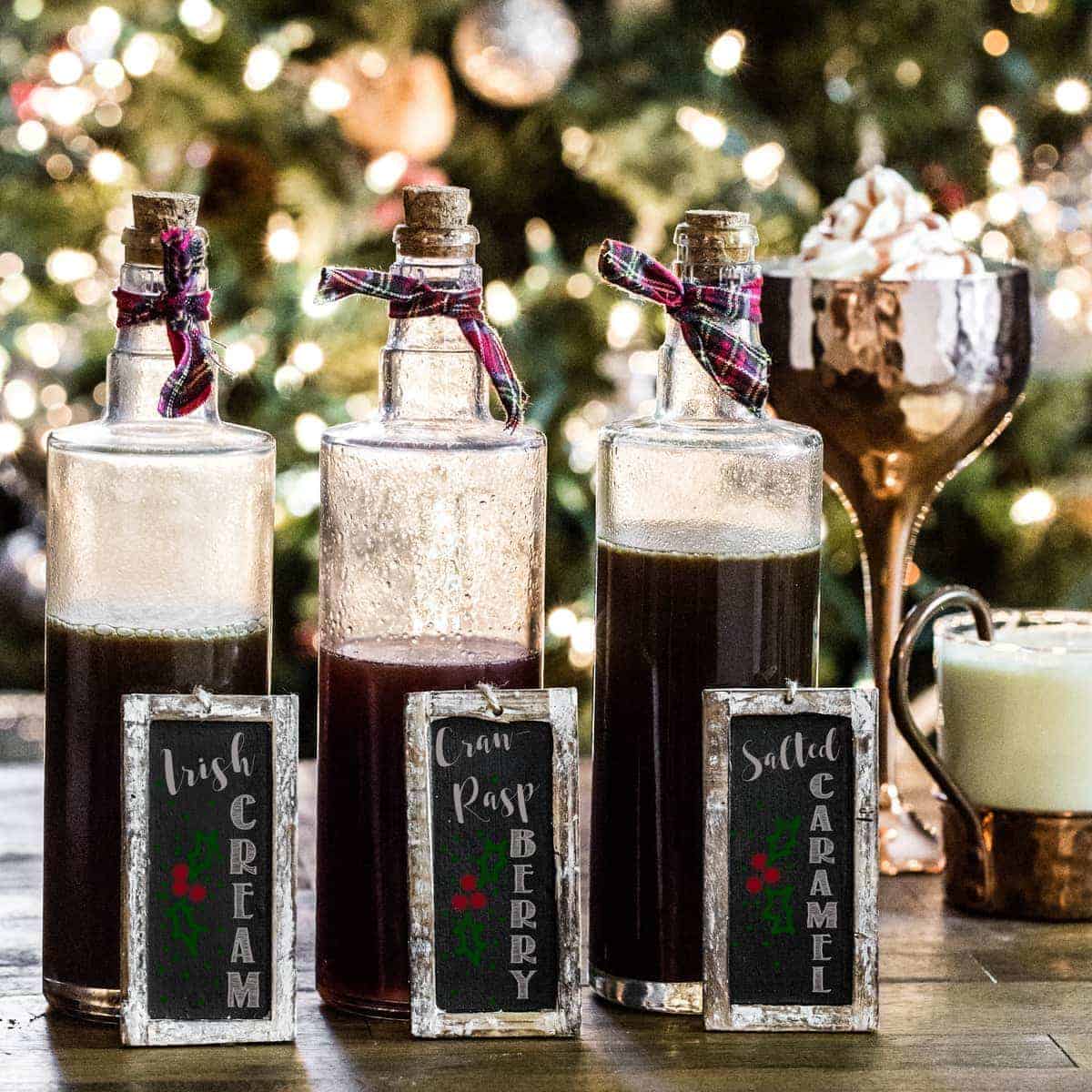 Three glass bottles of homemade syrup with chalkboard signs in front of a Christmas tree