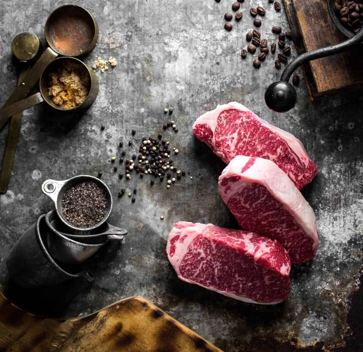 overhead ingredient shot of 3 strip steaks, brown sugar, peppercorns, a coffee grinder and a rosemary seasoning blend. 