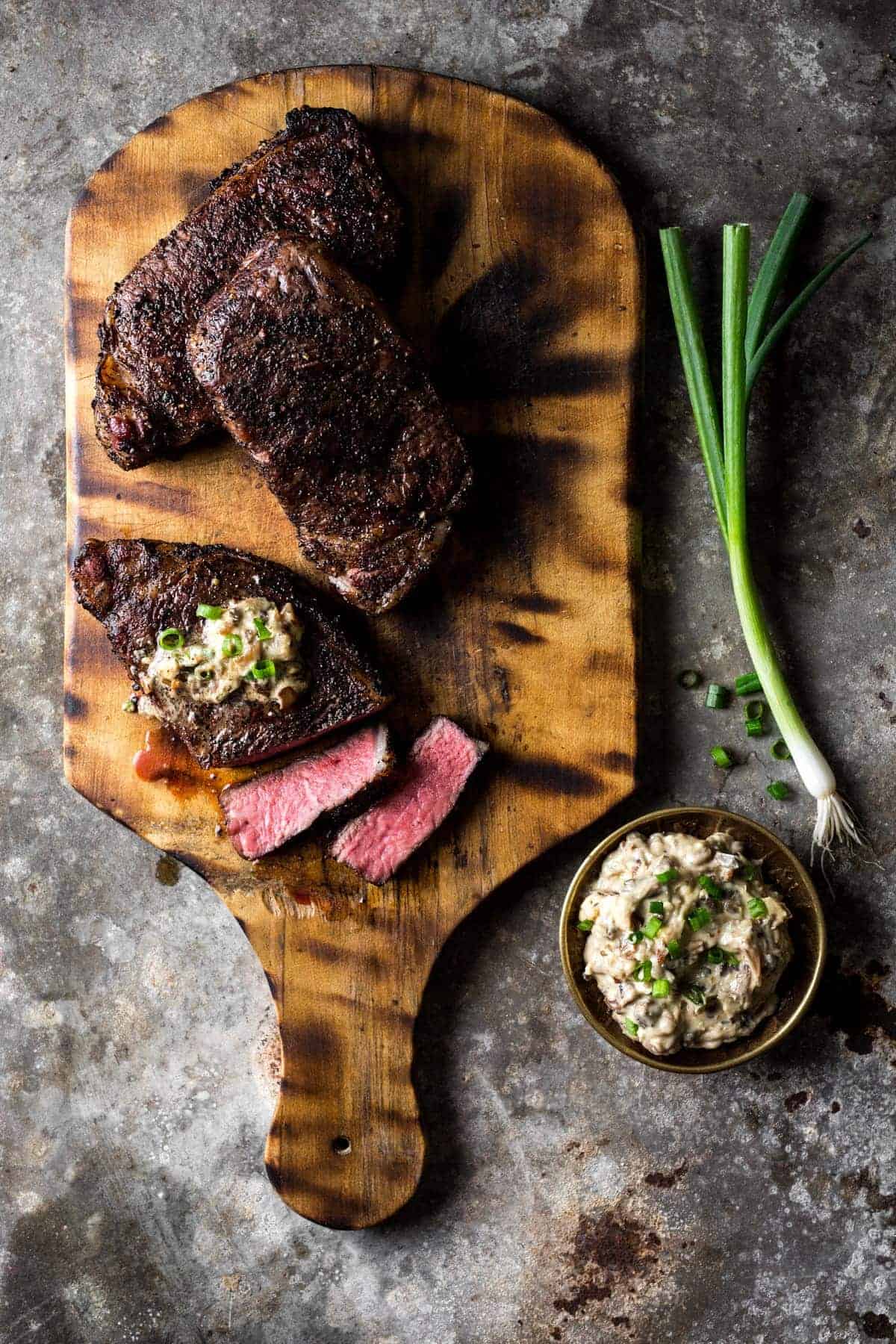 Fully cooked coffee crusted sous vide strip steak on a rustic cutting board. One steak sliced with blue cheese butter and green onion laying alongside