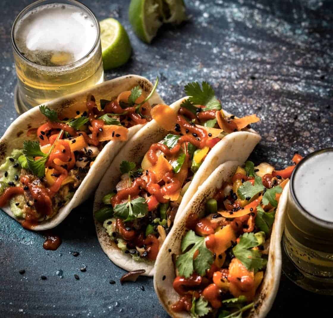 Three poke tuna tacos with bright vegetables and glasses of beer on a dark surface.