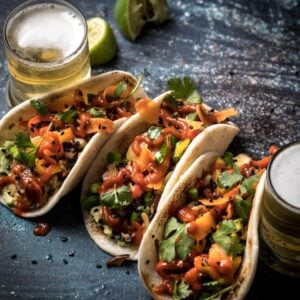 Closeup of three tuna poke tacos with glasses of beer on either side and a couple of squeezed limes.