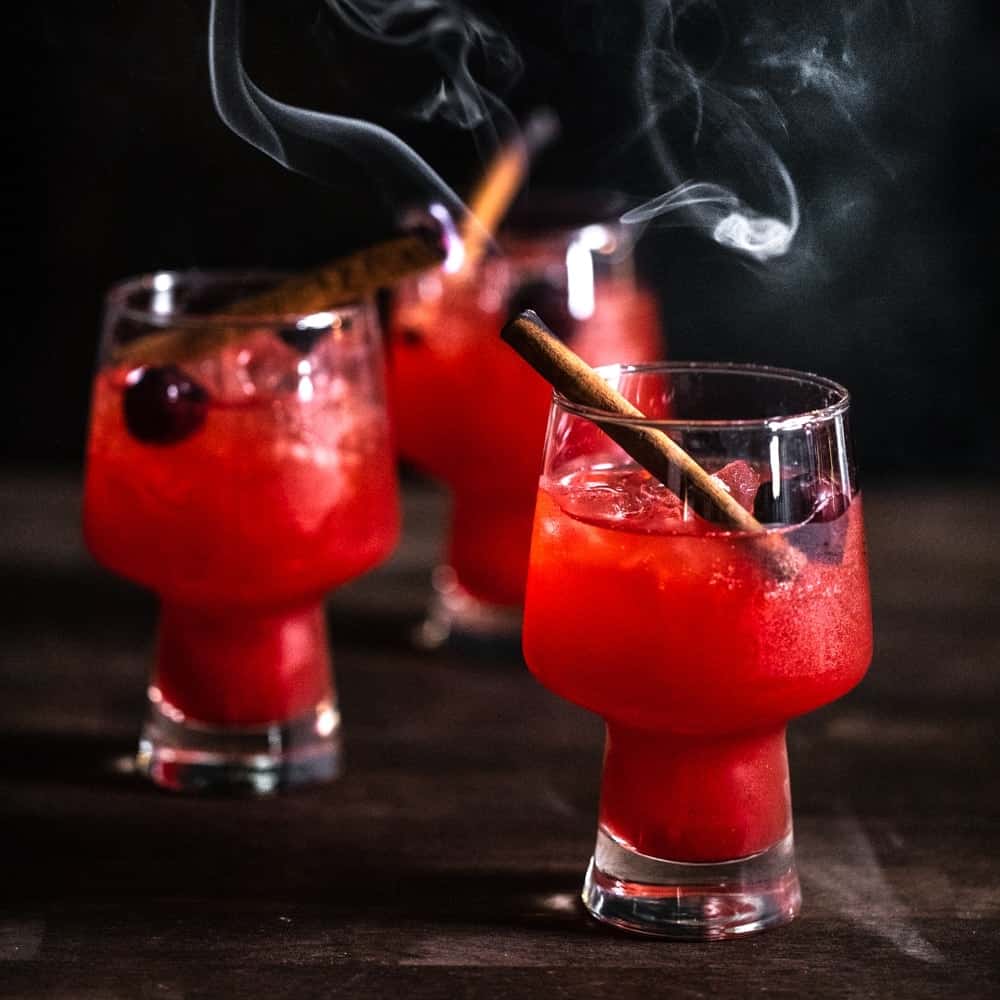 Trio of bright red Smoked apple old fashioneds with smoking cinnamon sticks