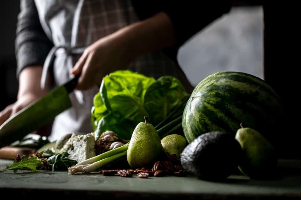 Jena chopping ingredients behind lettuce, watermelon, pears, onions, and avocado. 