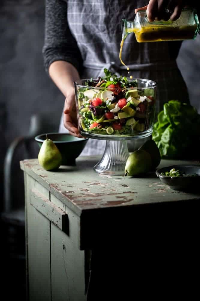 Pouring dressing into a glass serving bowl of melon avocado salad with a dark background.