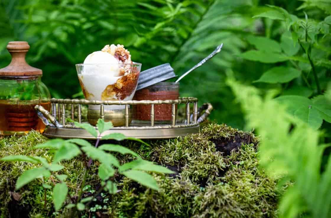 Gold tray with a frozen yogurt sundae, jar of strawberry rhubarb chia jam, and honey pot on a verdant stump. 