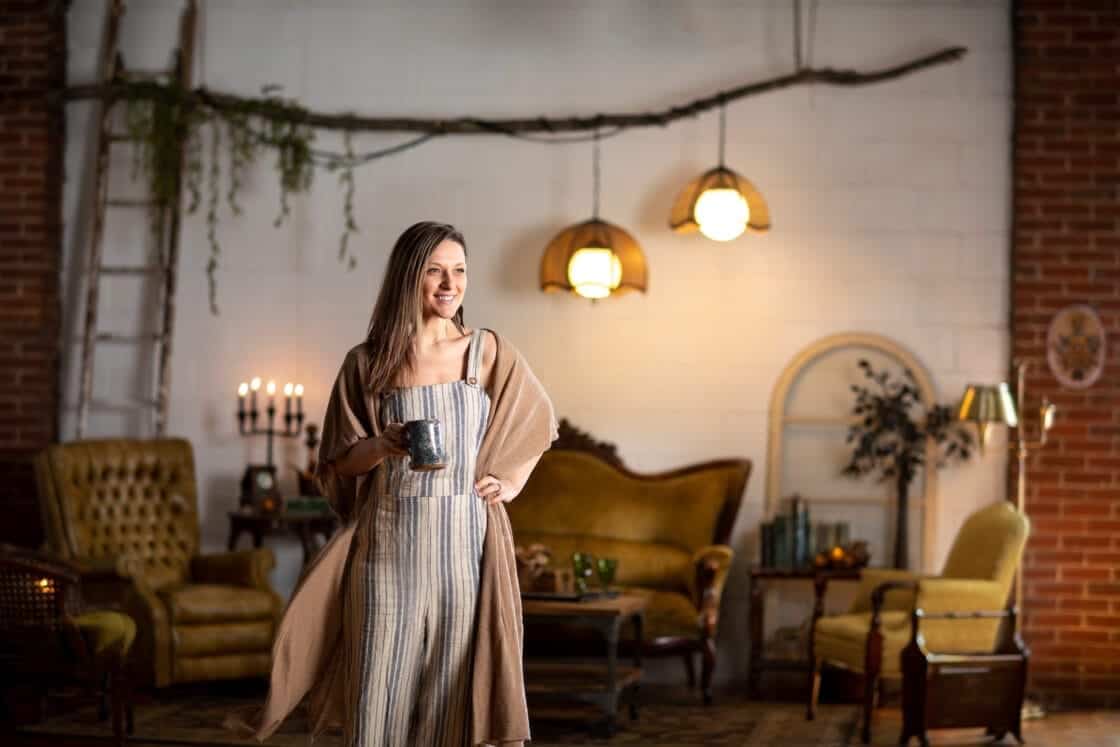 Female food photographer standing proudly in a cozy studio space with vintage props and furniture and exposed brick. 