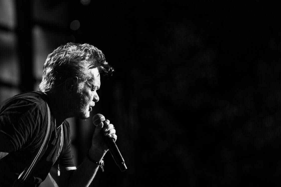 John Mellencamp singing into a microphone at Farm Aid with his profile dramatically back lit