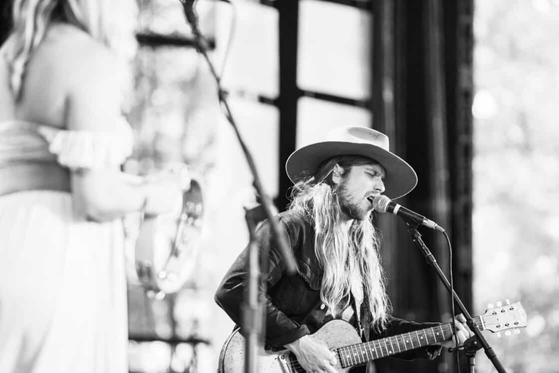 Lukas Nelson with long hair and a cowboy hat singing into a mic stand with his guitar at Farm Aid