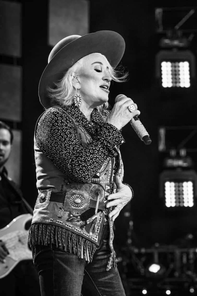 Tanya Tucket in a vest and cowboy hat singing into a microphone at Farm Aid. 