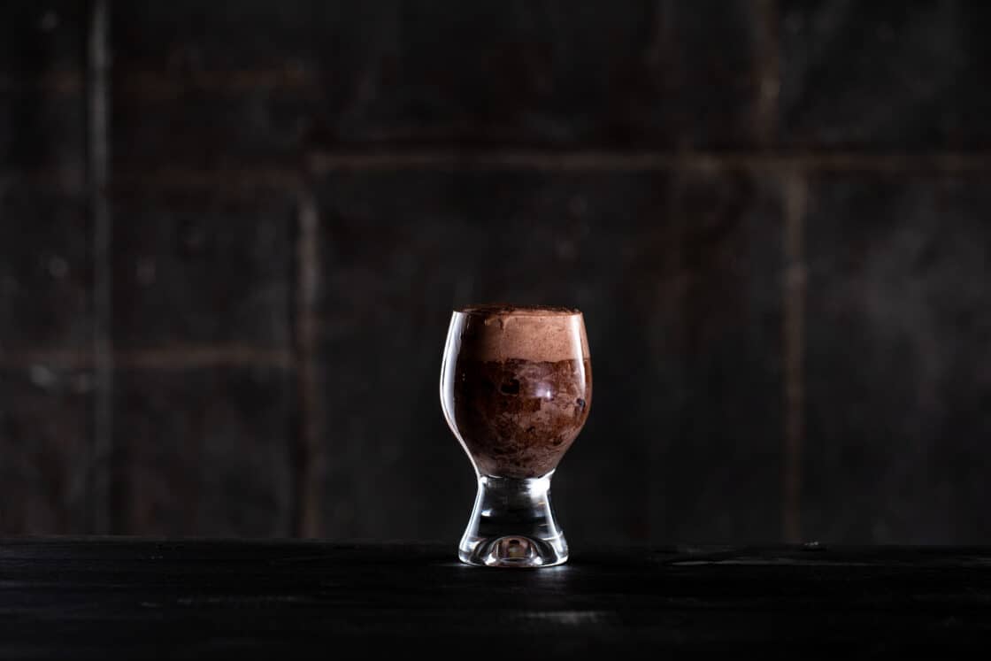 a single glass with a wide base filled with a brown beer float. Dark background with dramatic lighting. 