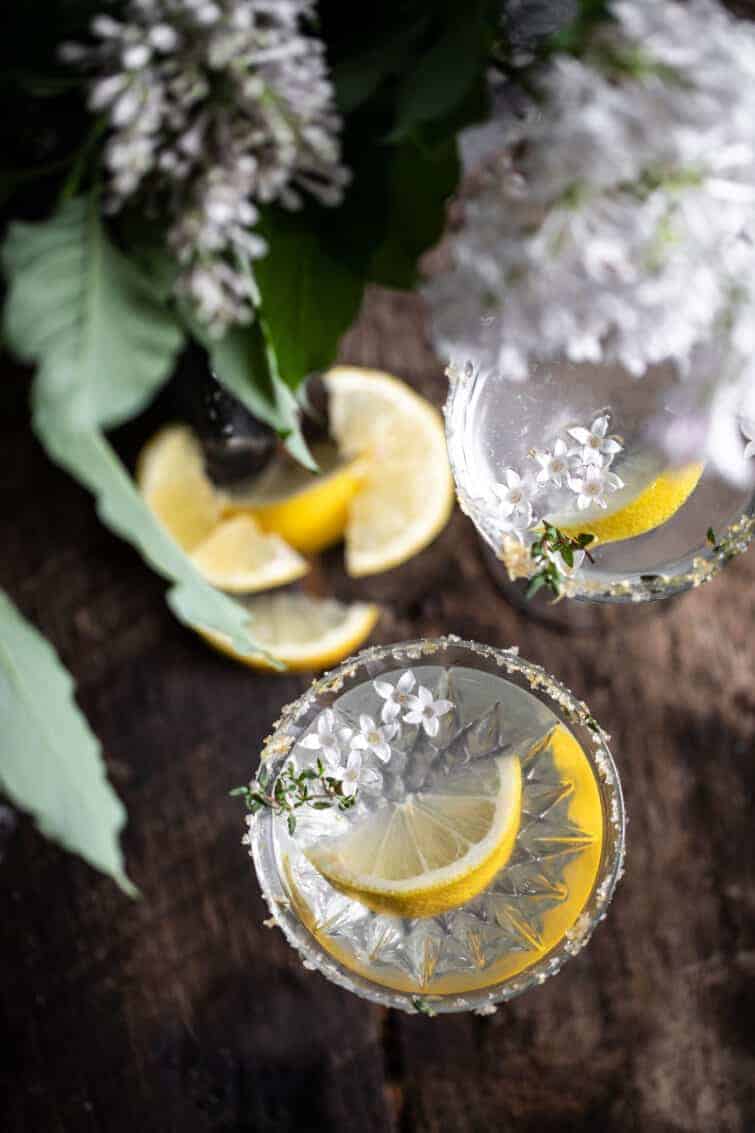 beautiful overhead garnished drink photography lemon drop martini in rustic wood setting with thyme lemon zest and lilac on the rim of the class recipe bordered by a bunch of white lilacs