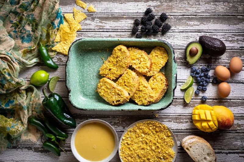 Process photo of layering the soaked and coated bread in the dish surrounded by fresh ingredients, sop, and crushed chips. 