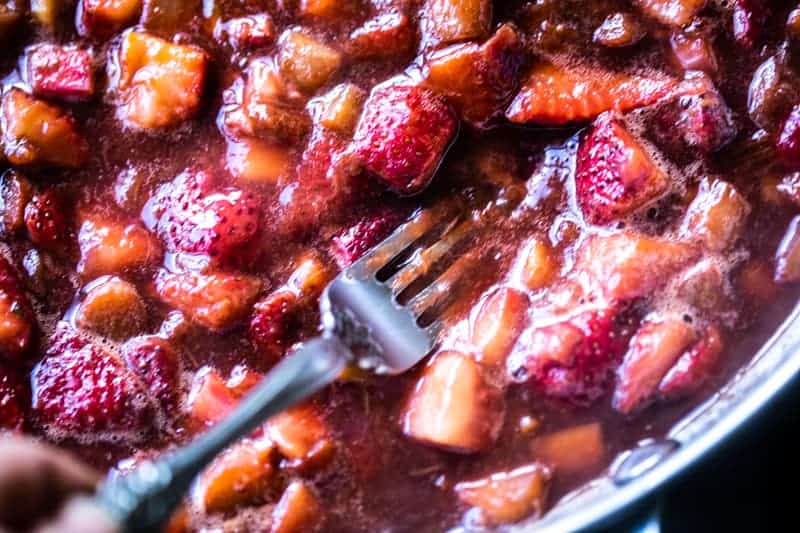 Closeup of cooked down strawberries and rhubarb with a fork mashing larger pieces. 