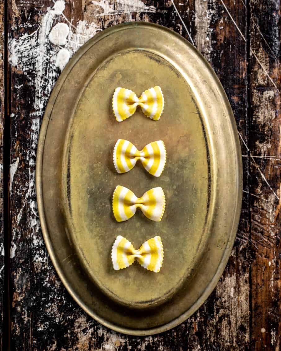 Four white and yellow striped bowtie pastas arranged on a antique platter and a rustic surface. 