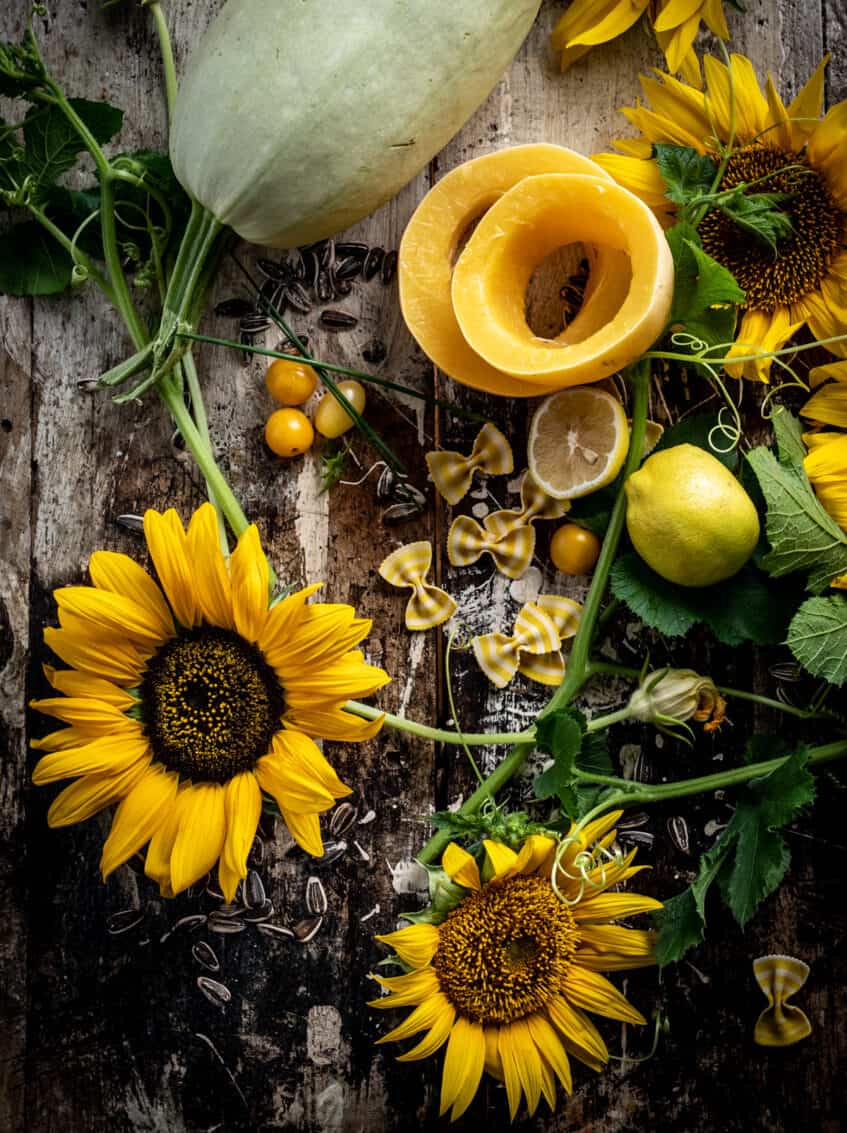 Overhead shot of artfully grouped ingredients and fresh sunflowers. Sunflower seeds, striped bowtie pasta, cut lemons and squash, and cherry tomatoes. 