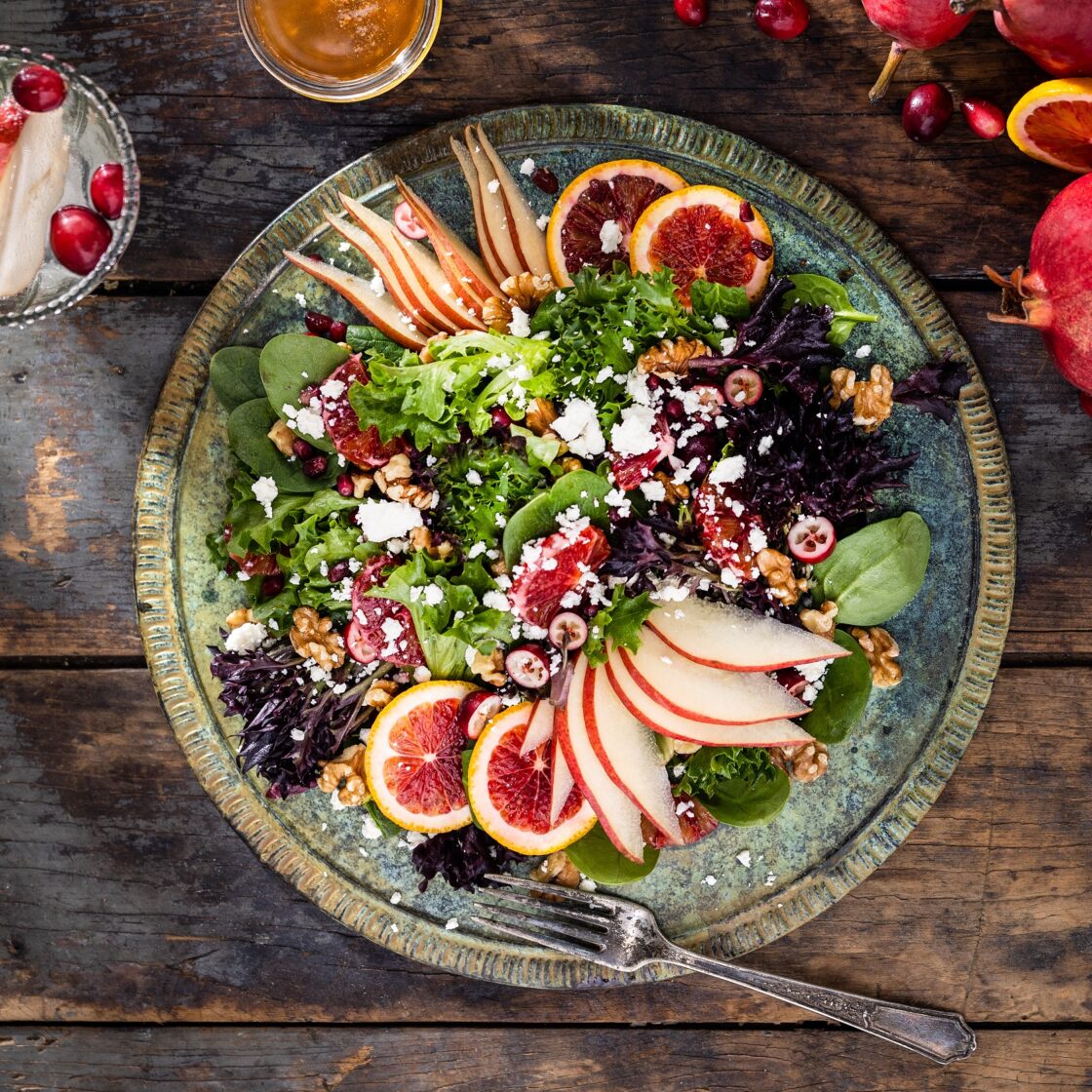Overhead view of a large plated green salad with red pear slices, blood oranges, cranberries, pomegranate, feta cheese crumbles, and walnuts