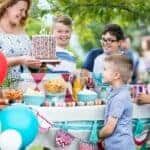A woman bring a sprinkled birthday cake to a picnic table with kids and balloons